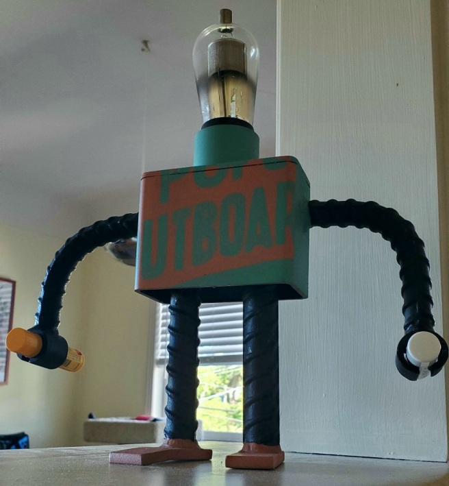 A wrought iron robot sculpture with a vacuum tube head is shown with two tubes of lip balm clamped into its hands.