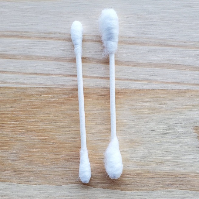 Two cotton swabs lay on a pine board. The cotton swab on the right appears to have been spun twice, making for much bigger, fluffy tips than usual.